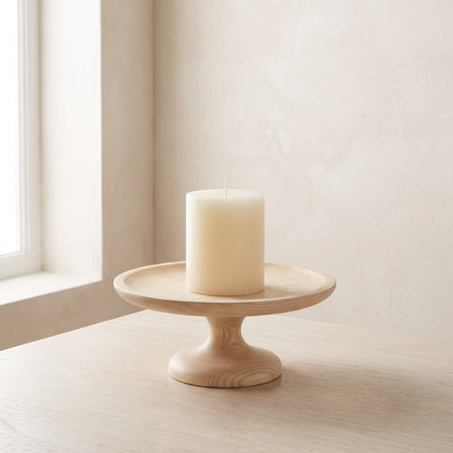 Pale ash wood pedestal candle holder with an ivory pillar candle on a light oak table by a window