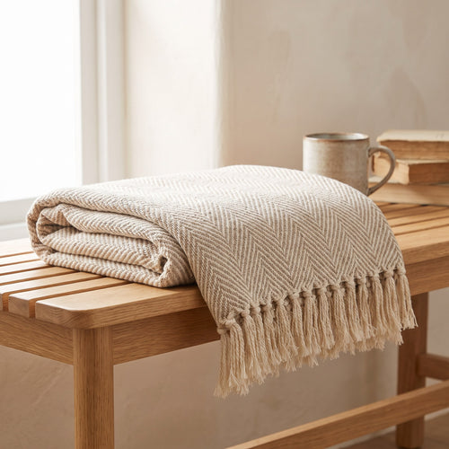 Oatmeal and cream herringbone cotton throw folded on an oak bench, knotted fringe visible