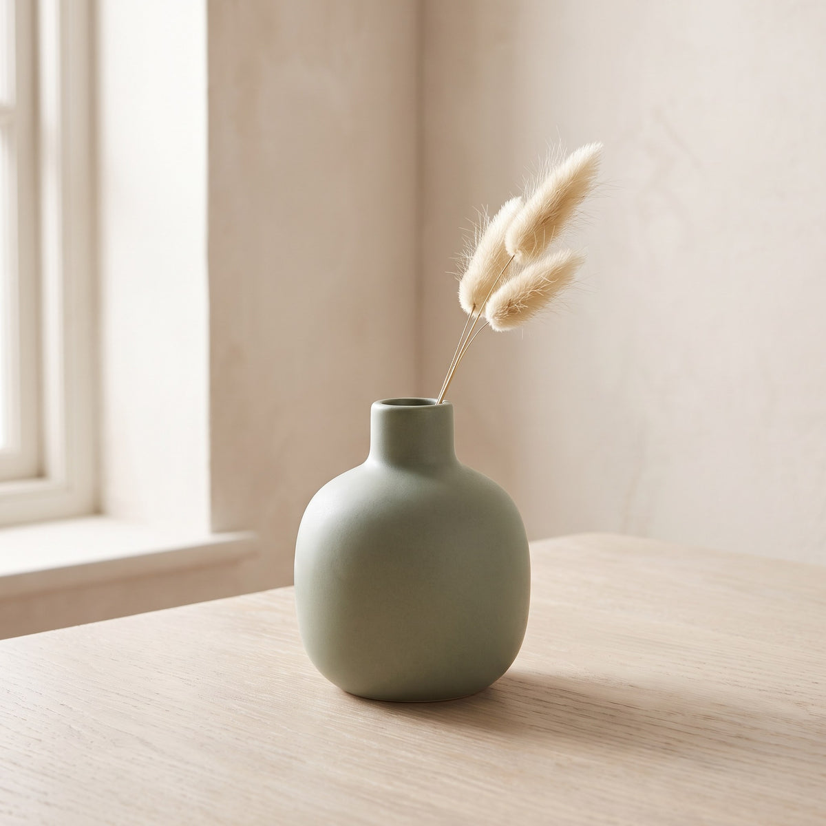 Small matte sage green stoneware bud vase with a single dried grass stem on a pale oak table