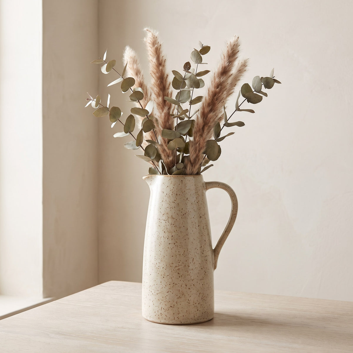 Speckled oatmeal stoneware jug vase with dried eucalyptus and pampas stems on a pale oak table