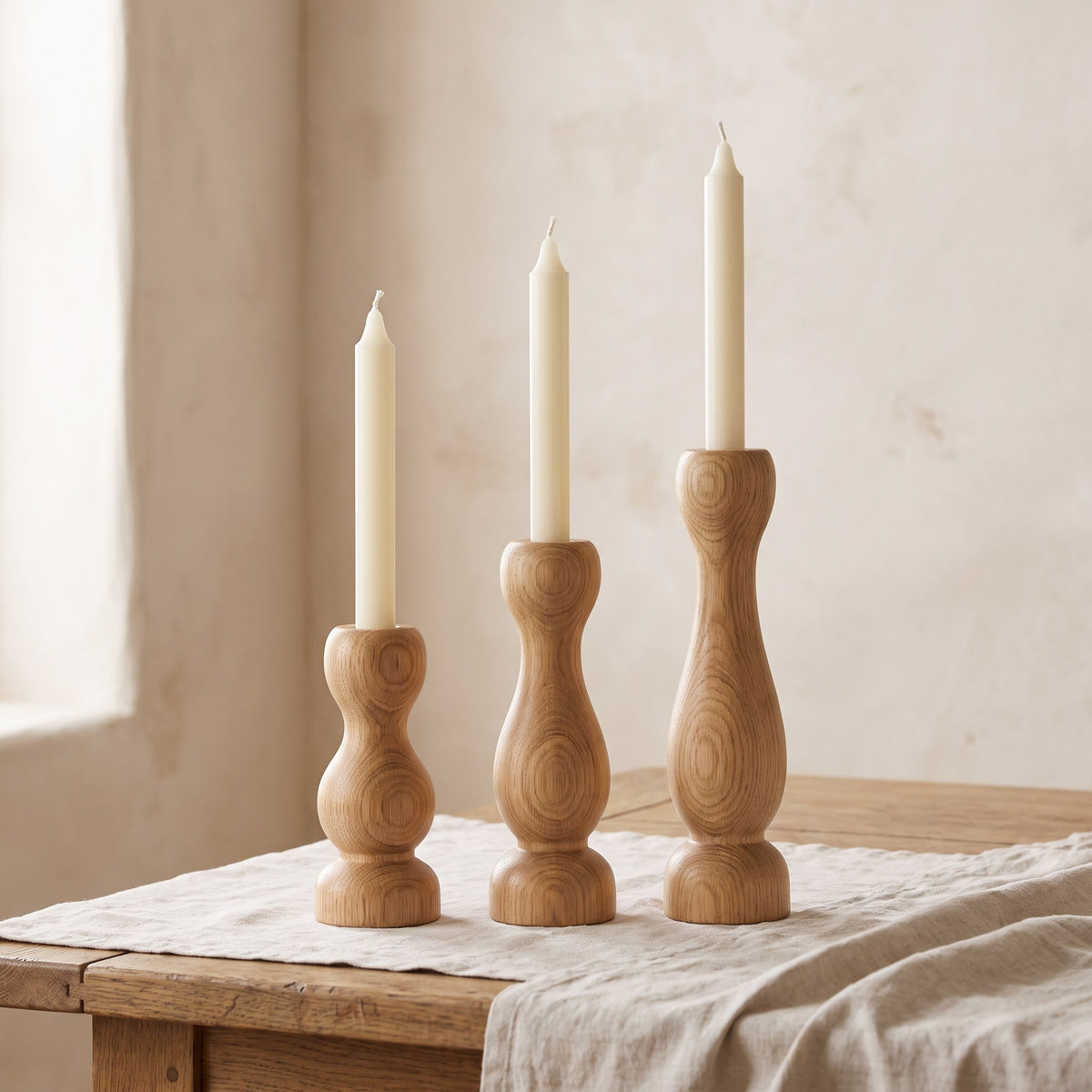 Three turned oak taper candle holders of different heights holding ivory dinner candles on a linen runner